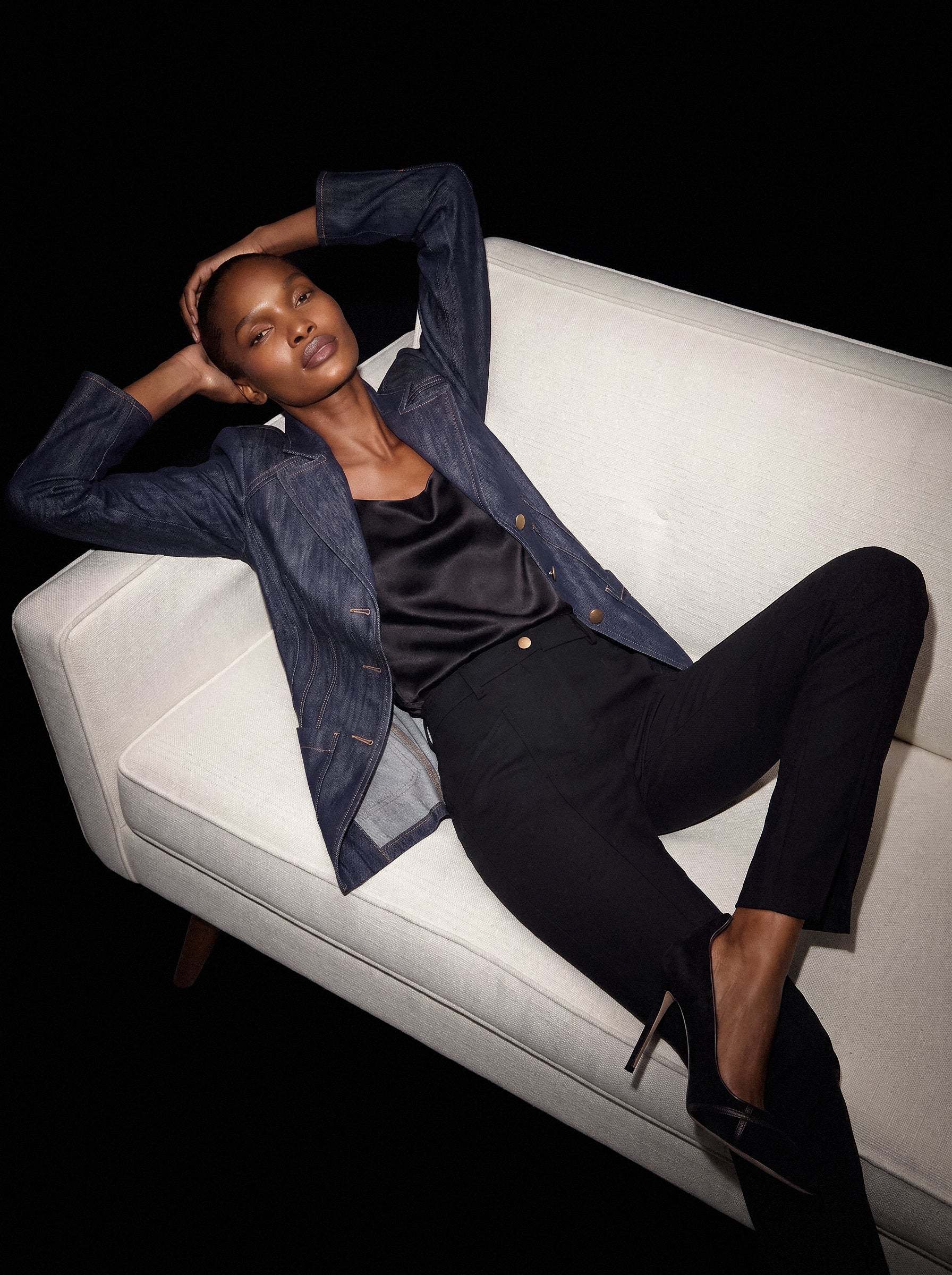 Model reclining on a cream sofa in an indigo denim jacket with contrast stitching and black silk top