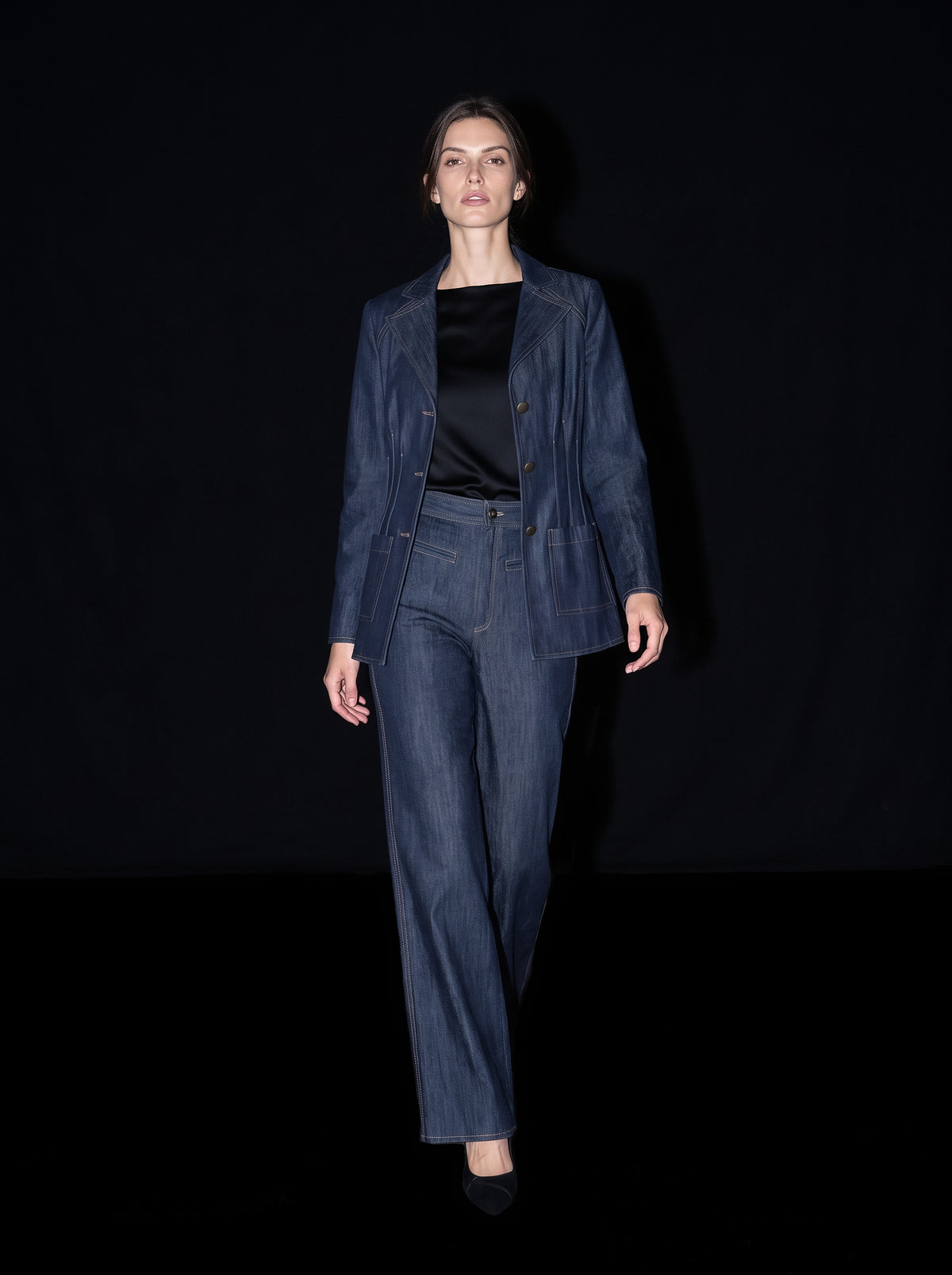 A model walks against a black background wearing a matching dark blue denim jacket and wide-leg jeans, styled with a black top and black shoes.