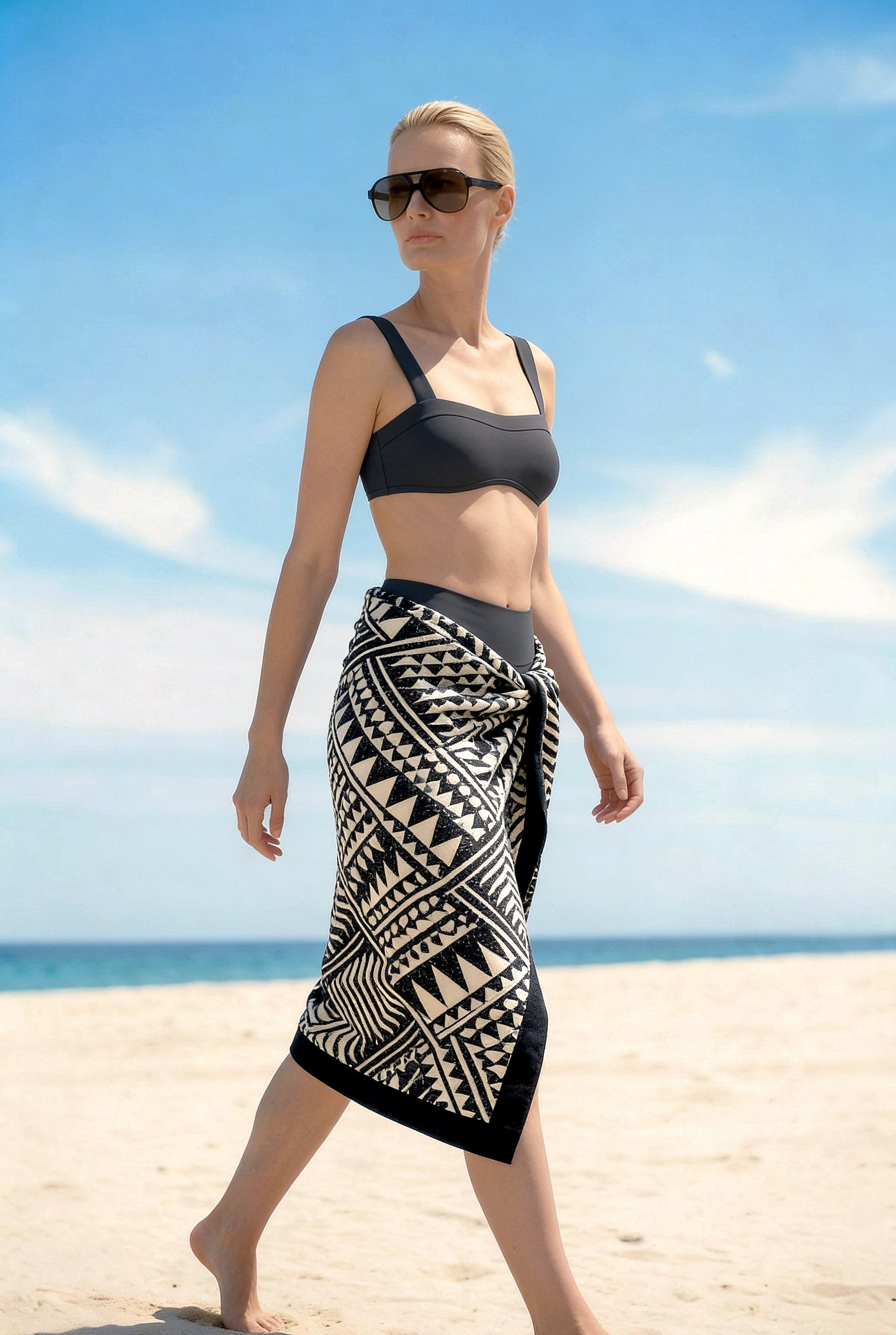 Woman in graphite swimsuit wrapped in a black and white geometric Isabella Kron beach towel, on a sunlit beach with a minimalist coastal backdrop