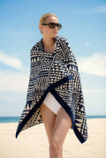 Woman in white swimsuit wrapped in a blue and white geometric Isabella Kron beach towel, styled as a luxury towel on a sunlit beach with a minimalist coastal backdrop
