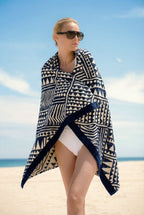 Woman in white swimsuit wrapped in a blue and white geometric Isabella Kron beach towel, styled as a luxury towel on a sunlit beach with a minimalist coastal backdrop