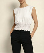 Woman wearing a white sleeveless top with a ruffled hem and black pants against a beige background
