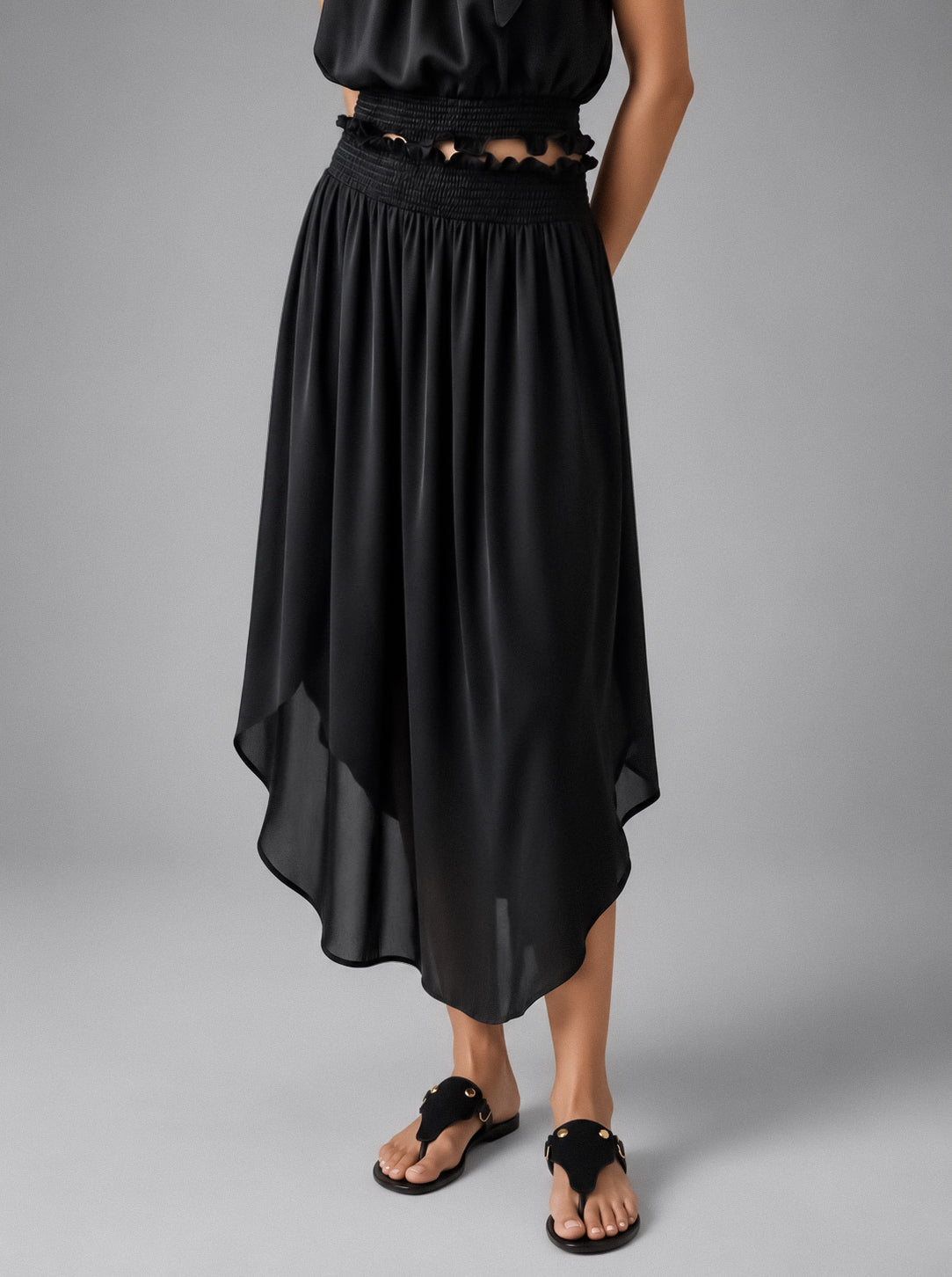A black high-low midi skirt with a smocked waistband and scalloped edge, shown on a model wearing black thong sandals against a gray studio background.