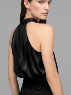 A close-up shot of a model from the back wearing a black satin halter top and a textured black skirt against a solid gray studio background.