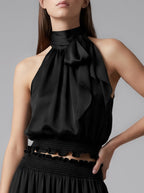 A model wearing a black halter-neck top with a large ruffled bow at the neck, paired with a matching black pleated skirt, against a solid gray studio background.