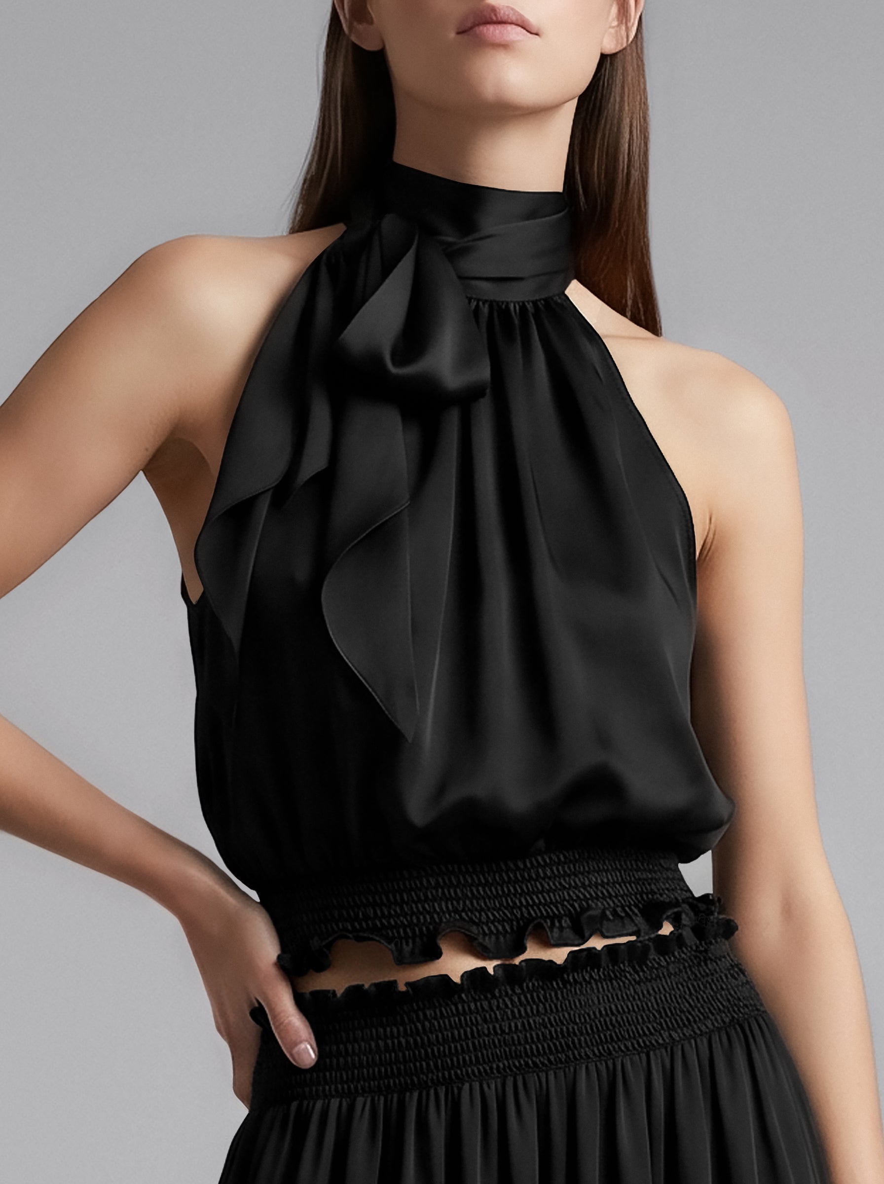 A close-up of a model wearing a black satin halter top with a large ruffled bow at the neck, paired with a black smocked skirt against a solid gray studio background.