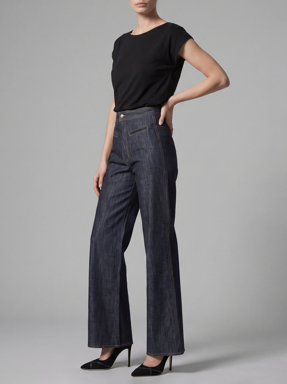 A model wearing dark blue wide-leg denim jeans, a black short-sleeve top, and black high heels, standing against a neutral gray studio background.
