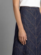 A close-up of a model wearing a dark indigo denim skirt with geometric chevron stitching, paired with a white top, against a solid gray studio background.