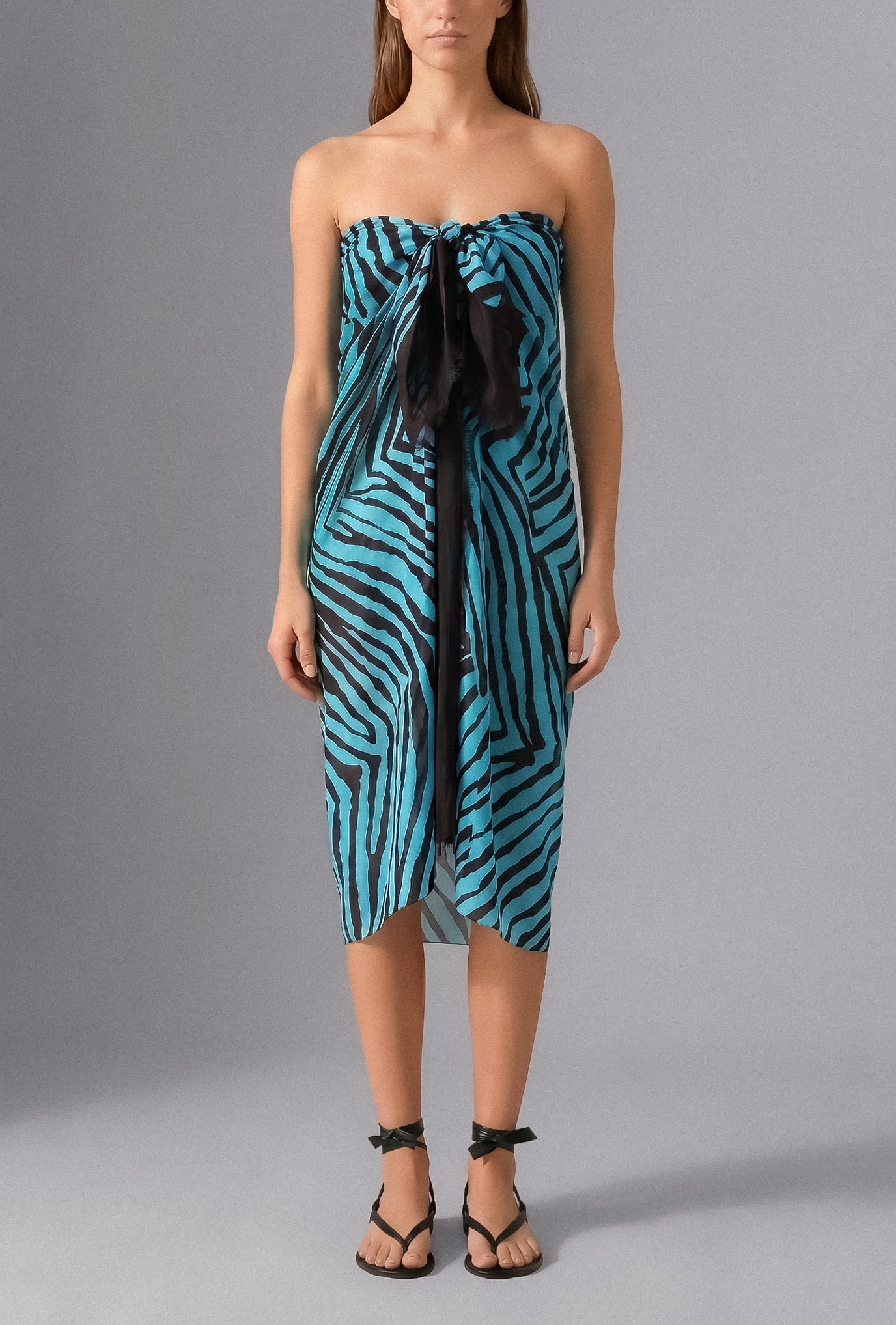 Woman wearing the Savana Pareo in turquoise and black graphic print styled as a lightweight luxury beach wrap over swimwear
